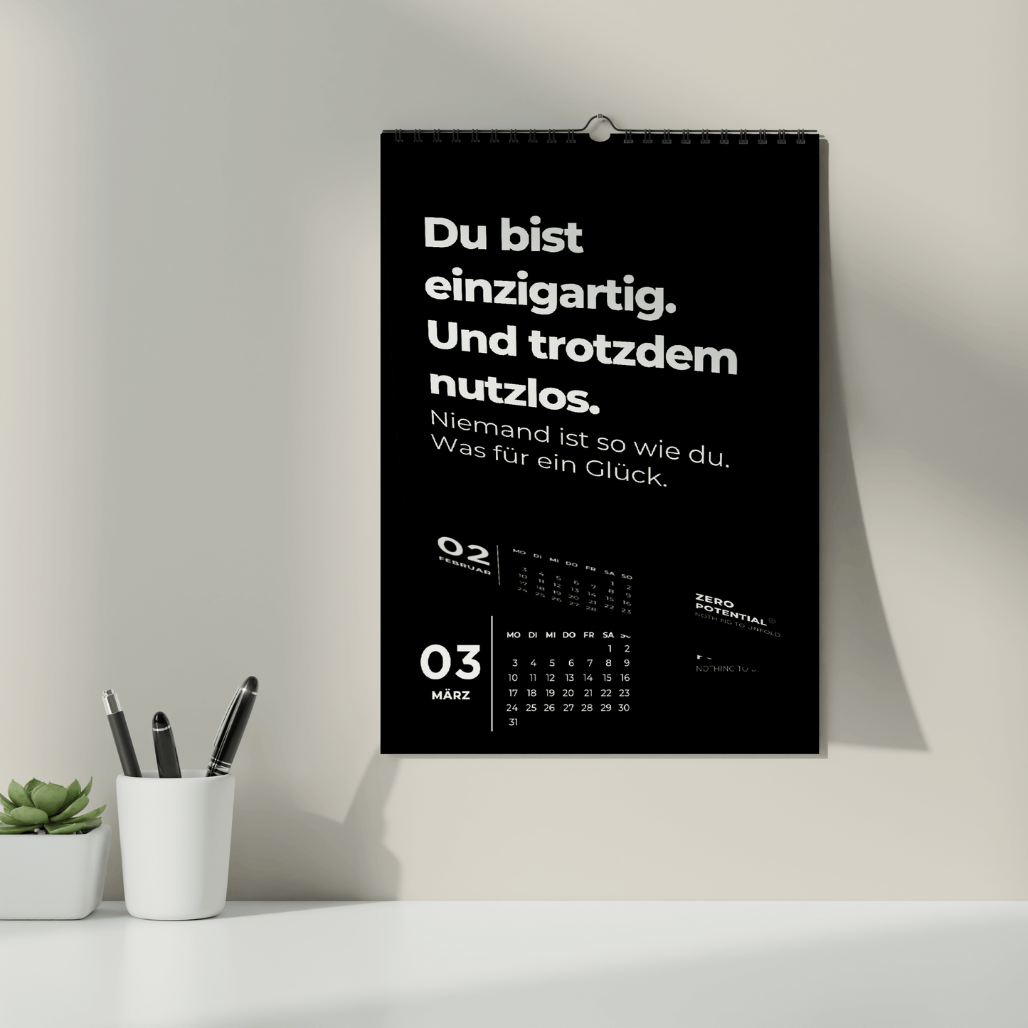 An der Wand hängt ein Kalender der Marke ZERO POTENTIAL, der stolz die deutsche Anti-Life-Coach-Botschaft zeigt: „Du bist einzigartig. Und trotzdem nutzlos.“ Dieser Demotivierender Wandkalender 2025 mit 12 unmotivierenden Sprüchen enthält ungeschönte Wahrheiten mit dem Datum 3. März, während unten auf dem weißen Schreibtisch eine Pflanze und Stifte stehen.